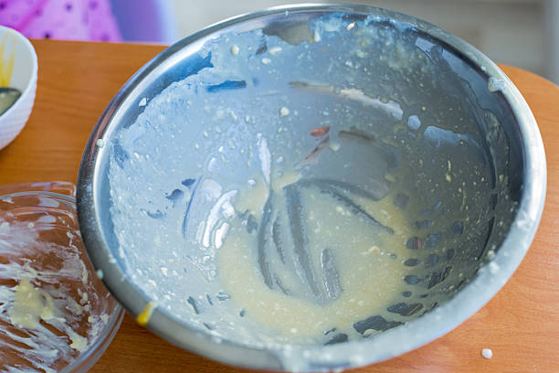 A little girl mixes batter and adds dried fruits while baking at home, showcasing a warm family cooking moment with bowls, whisk, and ingredients on a sunny kitchen table