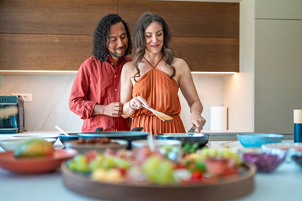 Close-up portrait of mid-adult interracial couple preparing food at kitchen counter. Man in red shirt and woman in orange dress smile while cooking with fresh ingredients.