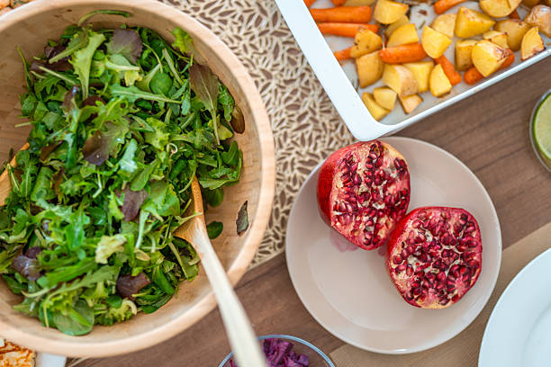 Freshly prepared dinner spread featuring a green leafy salad in a wooden bowl, sliced pomegranate, and roasted vegetables on a wooden table setting.