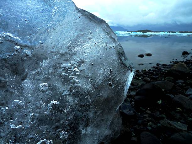Close-up of a textured ice block by a serene lake under a cloudy sky, showcasing natural beauty and tranquility.
