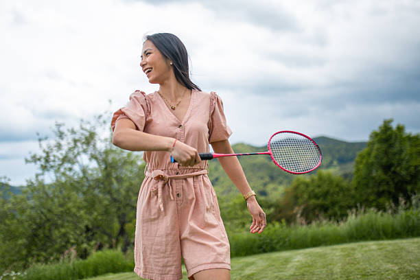 A cheerful young Asian woman in a pink dress plays badminton in a lush backyard under cloudy skies, surrounded by greenery and hills.