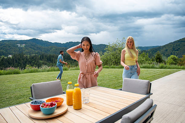 Lifestyle portrait of young women in casual summer attire sharing drinks and fresh fruit at wooden outdoor table, against dramatic mountain scenery and cloudy sky.