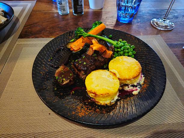 A gourmet dish featuring meat, vegetables, and potato gratin elegantly presented on a black plate on a wooden table.