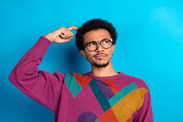 istock Young man with glasses scratching his head in thought against a bright blue background, wearing a colorful sweater 2248173637