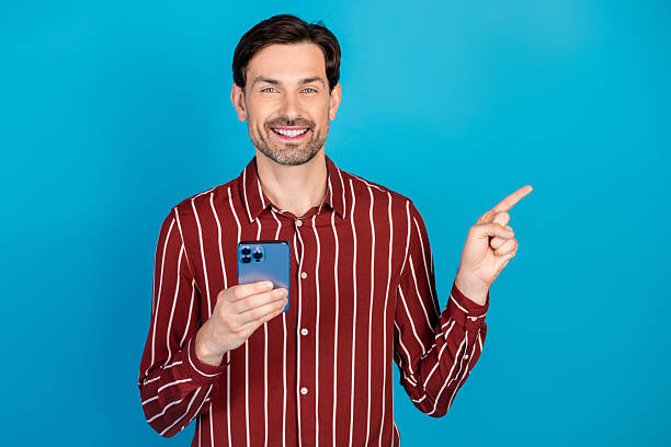 istock Young man with striped red shirt smiles while holding a smartphone and points toward the right against a blue studio background for lifestyle advertising and fashion stock photography 2248177142