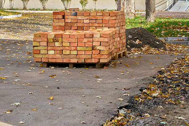 A massive heap of perforated and solid clay bricks forms a mountain-like pile in a dusty open area, surrounded by green trees in the background. The contrast between natural greenery and industrial material emphasizes the scale and rough texture of the construction debris. The image conveys abundance, durability, and outdoor storage.