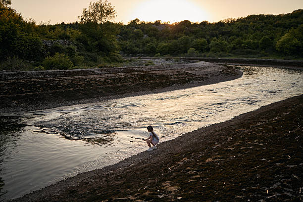 Young girl crouching by a river, playing with a stick at sunset