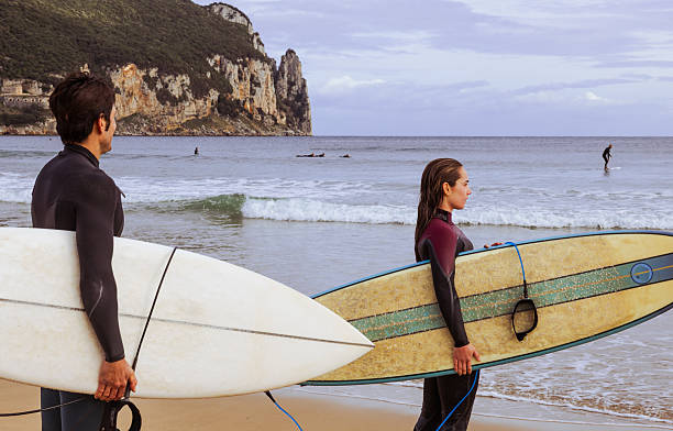 Two surfers stand side by side, watching the sea with boards in hand.