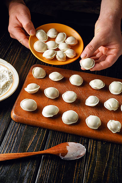 istock Hands are skillfully shaping dumplings on a wooden table. Nearby, a plate holds freshly formed dumplings while flour and a spoon rest beside them, indicating a cooking process in progress 2248199418