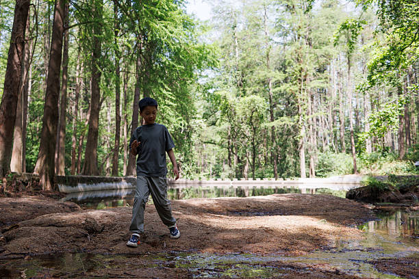 Young boy wearing a blue shirt and shorts walking barefoot on a sandy path, holding sticks in each hand. Background shows a park with trees