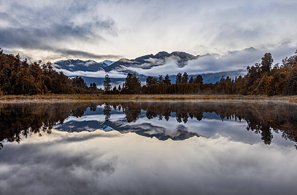 Misty clouds and forest surrounding Lake Matheson with perfect mountain reflection on calm water, South Island New Zealand