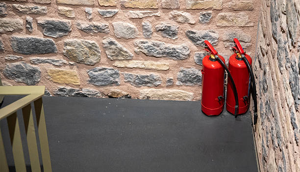 Two bright red fire extinguishers stand against a textured stone wall in a stairwell, ensuring fire safety and quick access during emergencies. The setting is well-lit and organized.