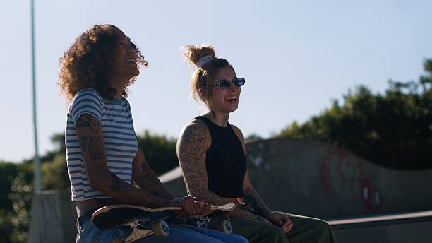 Two female skateboarders sitting together at a sunny outdoor skatepark, smiling and holding their skateboards.