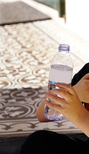 istock A boy holding a clear plastic water bottle with both hands, sitting on a patterned surface in bright natural light. 2248218878