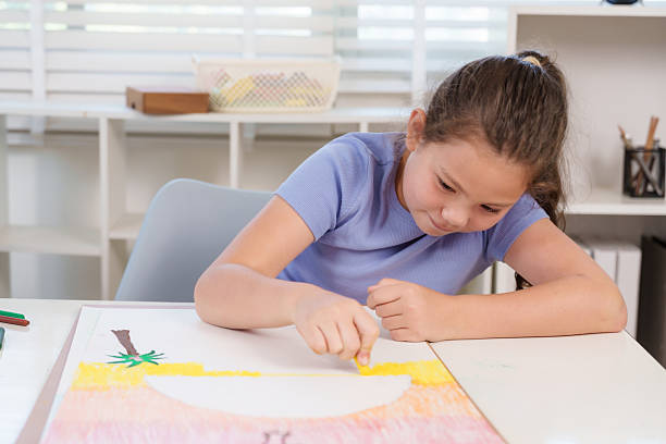 Caucasian elementary girl student creating artwork using yellow crayon on large paper at classroom desk representing creativity imagination childhood artistic expression educational learning