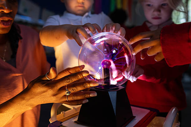 Close up selective focus shot of a group of young school children with their teacher standing around a plasma ball learning about it. Their faces are lit up. by the ball as they reach out to touch it. They are at a primary school in the North East of England.Videos are available similar to this shot.