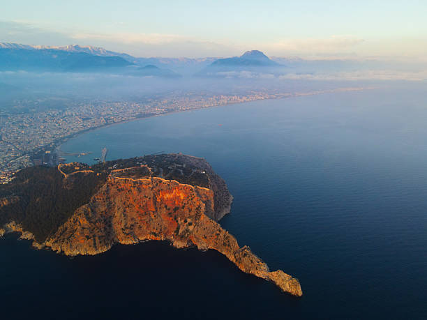 Dramatic aerial view of Alanya's rocky castle peninsula in the golden light of sunrise, with the city and hazy mountains visible along the coast