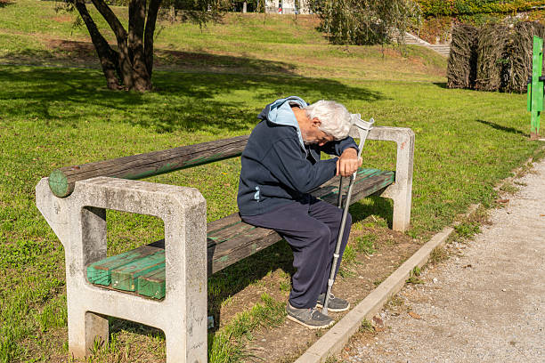 Senior man resting on park bench in city park on sunny day using crutches after hip operation.