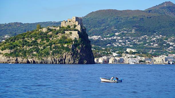 Ischia , Italy Oct 12,202 A Fishing boat floats by the coastline of the island of Ischia.   Aragonese Castle with the town of Ischia Ponte in the background