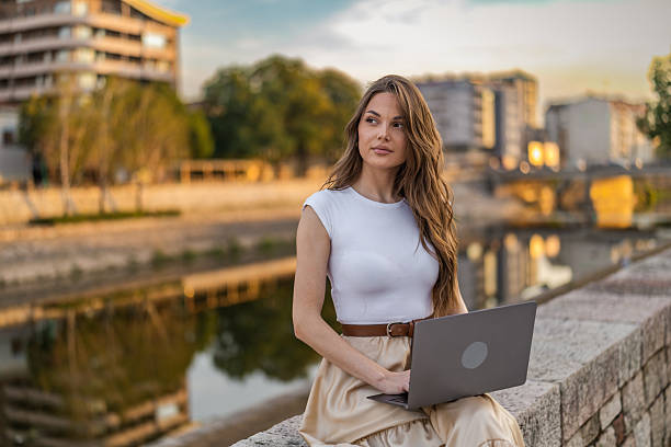 istock Beautiful girl with long hair freelancer work while sitting and using laptop on quayside in the city near the river. 2248235948