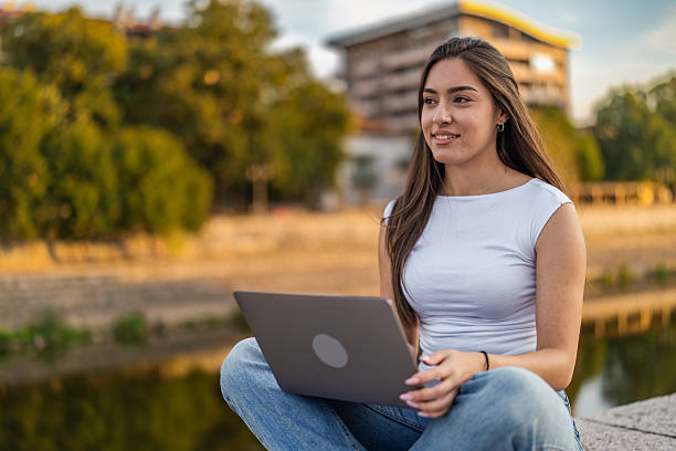 istock Beautiful girl with long hair freelancer work while sitting and using laptop on quayside in the city near the river. 2248236150