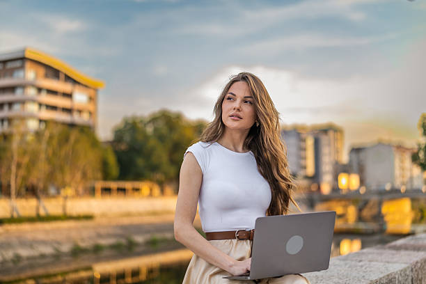 istock Beautiful girl with long hair freelancer work while sitting and using laptop on quayside in the city near the river. 2248236417