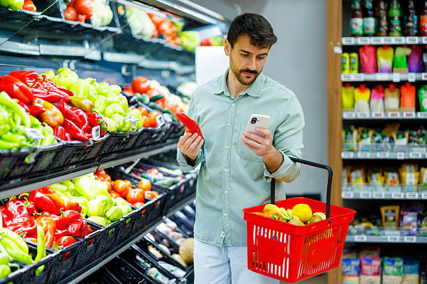 istock Man checking digital shopping list on smartphone while holding a red pepper and carrying a basket with fresh produce in a grocery store 2248237498