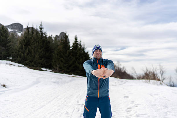 Adult man in activewear performing arm stretches and warming up for outdoor exercise on a snow-covered mountain, surrounded by pine trees under a cloudy sky
