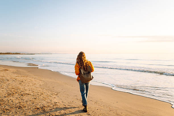istock A smiling woman in a bright yellow jacket strolls along a sandy beach at sunset. 2248249488
