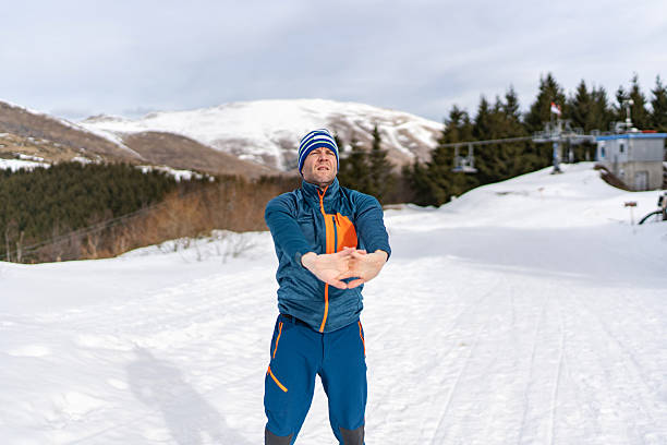 Middle-aged man stretching and warming up on a snowy mountain slope at a ski resort, preparing for a winter workout or ski run amid rugged alpine scenery