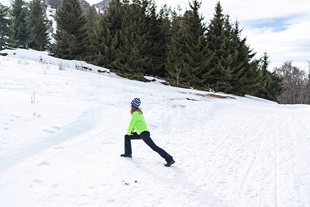 Young woman doing a deep lunge stretch on a snow-covered mountain trail, staying active and focused on fitness amid pine trees and clear blue winter sky, bundled in warm gear