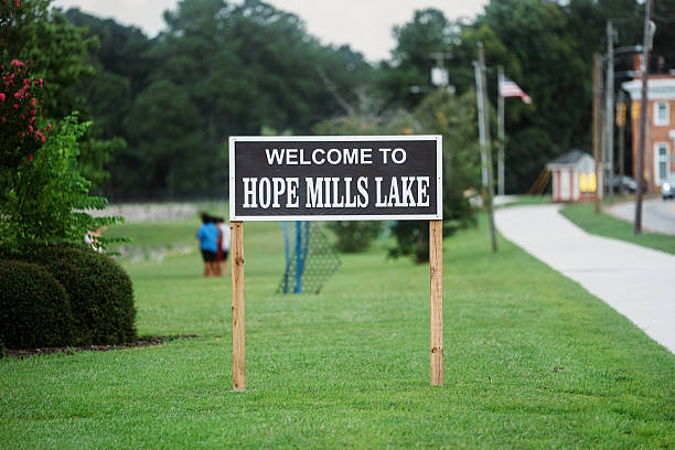 A wooden sign reading “Welcome to Hope Mills Lake” stands along a grassy lawn near the lakeside walkway, marking the entrance to the community park on a calm summer day.