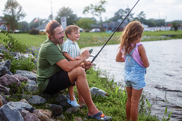 A three-year-old Eurasian boy giggles with excitement as his uncle sits on the rocky shoreline and teaches him how to fish during an easy-going multi-family summer trip to the lake.
