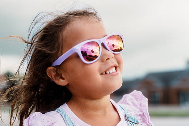 A young Eurasian girl smiles softly as her purple sunglasses reflect the sky and street signs during a breezy summer afternoon stroll.