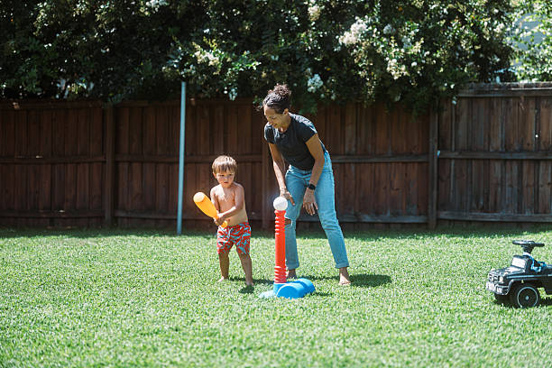 A Eurasian mom spends quality time helping her toddler learn to swing a bat during a playful backyard t-ball session on a warm summer day.