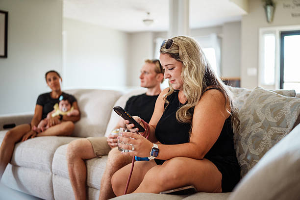 A woman scrolls through her phone and sips a drink while sitting in the bright and welcoming living room of neighborhood friends during a casual afternoon visit.