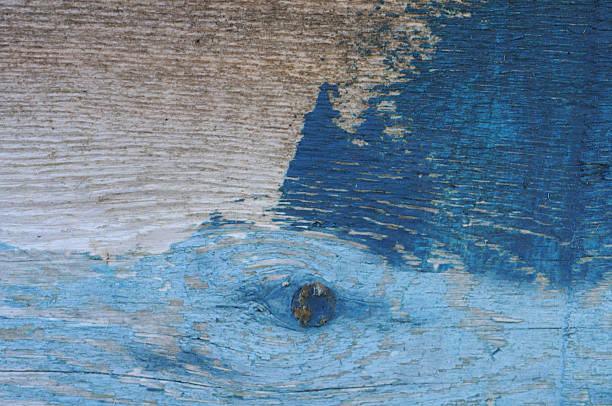 A textured wooden surface displays a mix of faded blue paint and exposed natural wood grain. The paint is peeling and weathered, revealing underlying patterns and knots in the wood, including a prominent knot in the center. The wood's texture is enhanced by horizontal grain lines, creating an abstract and rustic appearance. The wear and color variation suggest age and exposure to elements.