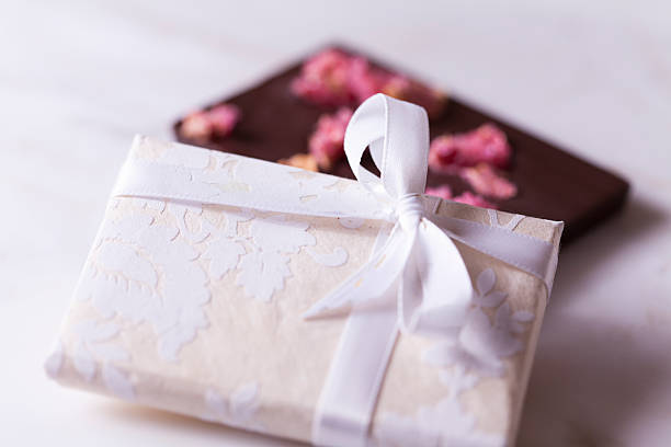 A small laced gift box with a white ribbon tied in a knot, lying on a rectangular piece of dark chocolate with dried roses on white.