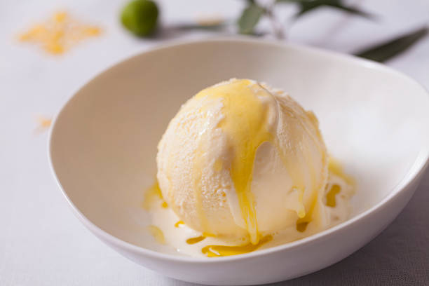 A large scoop of vanilla ice cream with yellow cream  in a small white bowl on a white table.