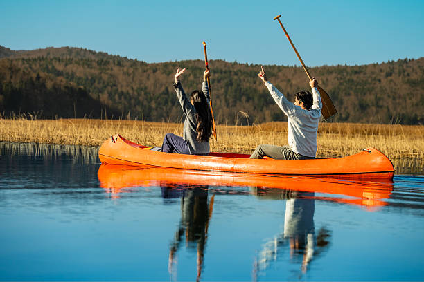 Two individuals in an orange canoe on a serene lake, holding paddles aloft in celebration. Golden reeds and distant hills surround them, embodying joy and freedom amidst nature's tranquility. The clear water reflection enhances the peaceful ambiance.