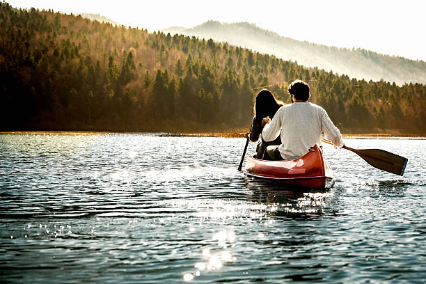 A couple paddles a vibrant orange canoe on a serene lake, surrounded by lush, forested hills. The clear blue sky and calm water reflect the peaceful outdoor setting, capturing the essence of adventure and tranquility in nature.