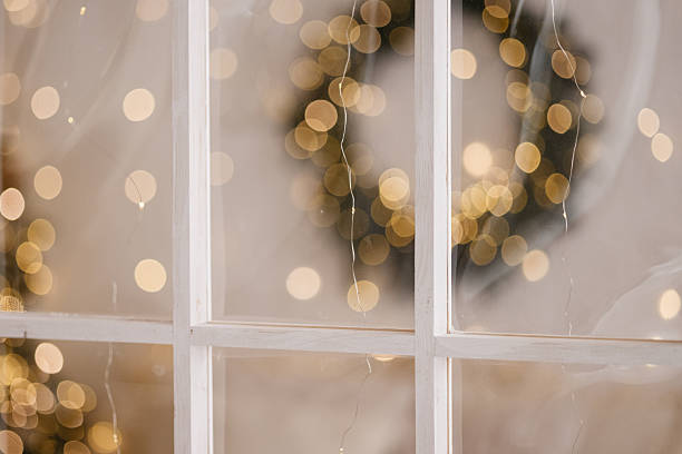 White wooden window frame with glass in an indoor festive setting, showing blurred warm lights and a wreath detail. Suitable for holiday themes, decoration concepts, celebration visuals.