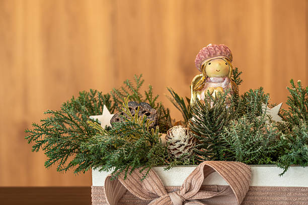 A festive close-up of a Christmas angel figurine placed among pine branches, frosted cones, wooden stars, and greenery. The decoration is arranged in a cream-colored box wrapped with a pink ribbon, set against a warm wooden background.