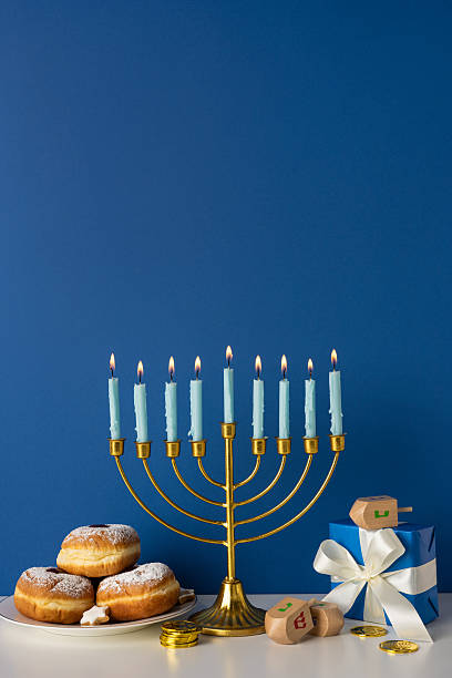 A vertical festive Hanukkah scene with a lit menorah, soft candle glow, donuts and coins, and wrapped gifts on a blue backdrop. Warm, celebratory mood suitable for holiday greetings stock photo