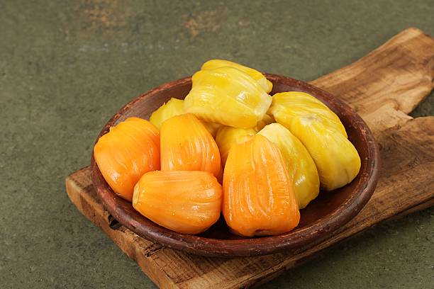 Close up of vibrant yellow jackfruit pods arranged in a wooden bowl on a rustic surface, showcasing fresh tropical fruit texture and color.