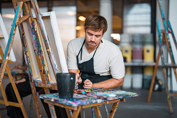 Mid adult Caucasian male painter in a bright art studio, wearing an apron, deeply engaged in creating artwork with vibrant colors and tools on a wooden easel. The studio reflects a creative and inspiring atmosphere.