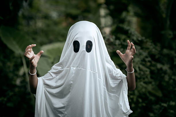 A young boy dressed as a ghost for Halloween wears a white sheet with two black eye holes. He raises both arms in a spooky gesture, surrounded by dark green foliage
