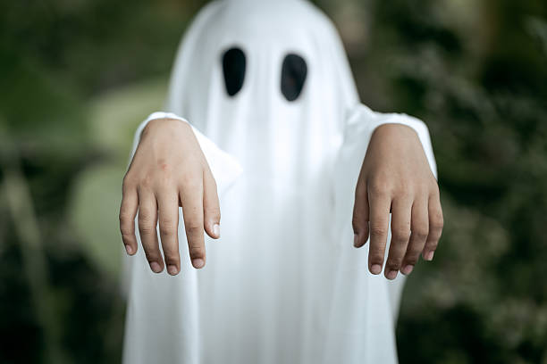 istock A young boy dressed as a ghost for Halloween wears a white sheet with two black eye holes. He raises both arms in a spooky gesture, surrounded by dark green foliage 2248300617