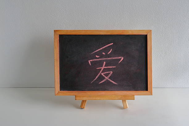 Chinese character means love, is written on chalkboard resting on white wooden table against white concrete wall. The entire setup creating a simple and minimalist composition.