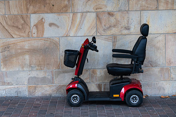 A red red senior citizen mobility scooter stands still next to a textured stone wall. The scooter is ready for use, featuring a comfortable seat and basket for convenience.